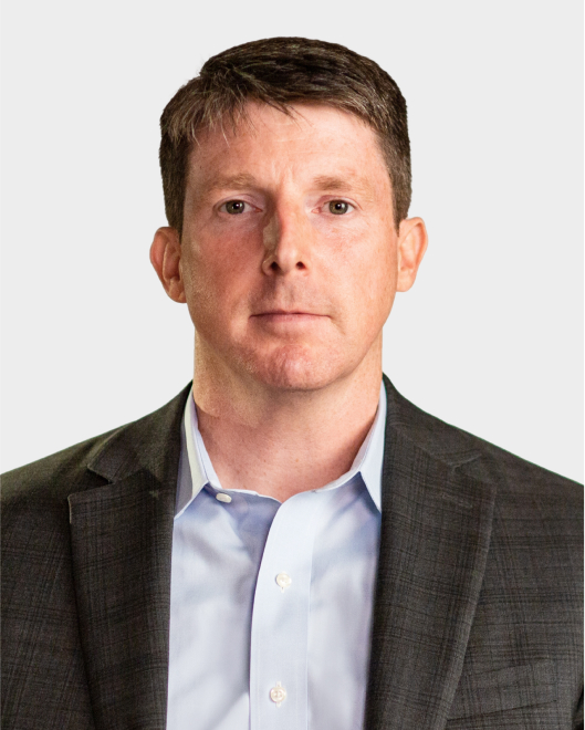 A man with short brown hair wearing a gray suit jacket and light blue shirt, facing forward with a neutral expression against a plain background.