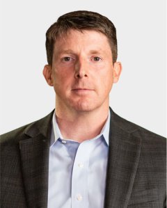 A man with short brown hair wearing a gray suit jacket and light blue shirt, facing forward with a neutral expression against a plain background.
