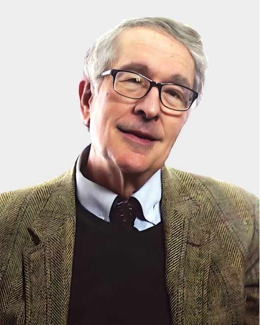 An older man with gray hair and glasses is wearing a brown herringbone jacket, a collared shirt, and a dark sweater, posing against a plain light background.