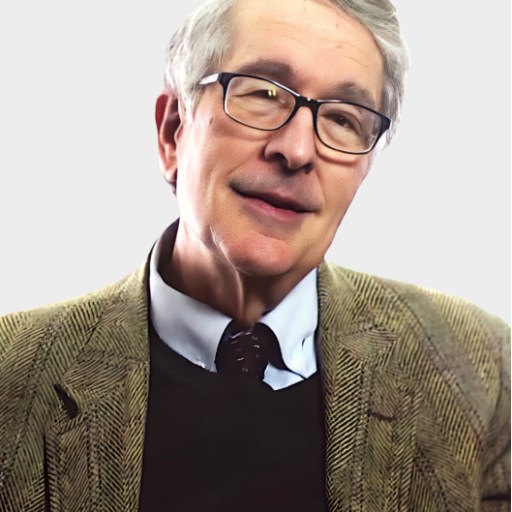 An older man with gray hair and glasses is wearing a brown herringbone jacket, a collared shirt, and a dark sweater, posing against a plain light background.