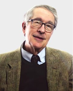 An older man with gray hair and glasses is wearing a brown herringbone jacket, a collared shirt, and a dark sweater, posing against a plain light background.