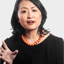 A woman with medium-length black hair wears a black top and an orange beaded necklace, gesturing with her left hand while speaking against a plain light background.