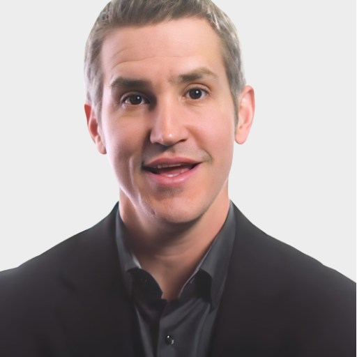 A man with short, light brown hair wearing a black suit and dark shirt is speaking, set against a plain light background.
