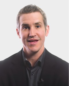 A man with short, light brown hair wearing a black suit and dark shirt is speaking, set against a plain light background.