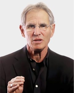 An older man with gray hair and glasses, wearing a black jacket and shirt, gestures with his right hand against a plain light background.