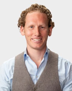 A man with curly hair, wearing a light blue dress shirt and a grey vest, smiles at the camera against a plain background.