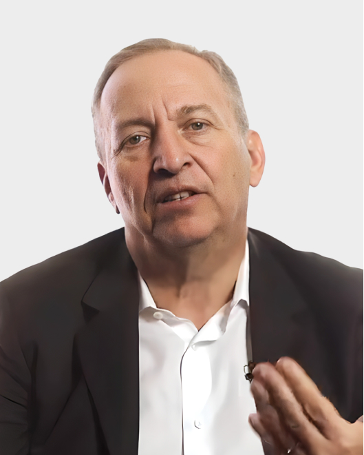 A middle-aged man in a dark suit and white shirt speaks, gesturing with his hand, against a plain light background.