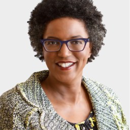 A woman with short curly hair and glasses smiles at the camera. She is wearing a patterned jacket over a patterned top, in front of a plain light background.