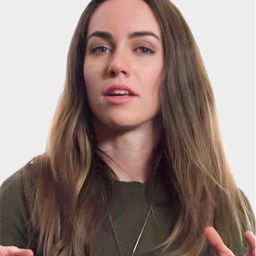A woman with long brown hair and a neutral expression gestures with her hands. She is wearing a dark top and a pendant necklace against a plain background.
