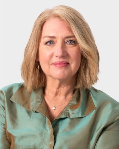 A middle-aged woman with shoulder-length blonde hair wearing a light teal satin blouse, looking at the camera against a plain background.