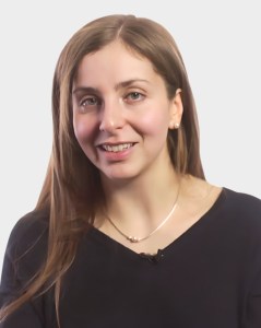 A woman with long brown hair, wearing a black top and a necklace, looks at the camera and smiles against a plain light background.