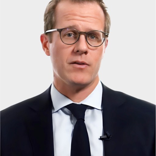 A man wearing glasses, a dark suit, white shirt, and dark tie stands against a plain light background, looking directly at the camera.