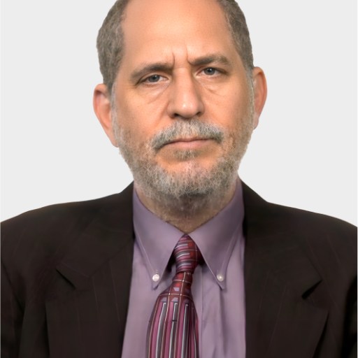 A middle-aged man with short gray hair and beard wears a brown suit, mauve shirt, and striped tie, facing the camera against a plain light background.
