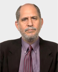 A middle-aged man with short gray hair and beard wears a brown suit, mauve shirt, and striped tie, facing the camera against a plain light background.