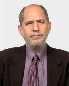 A middle-aged man with short gray hair and beard wears a brown suit, mauve shirt, and striped tie, facing the camera against a plain light background.