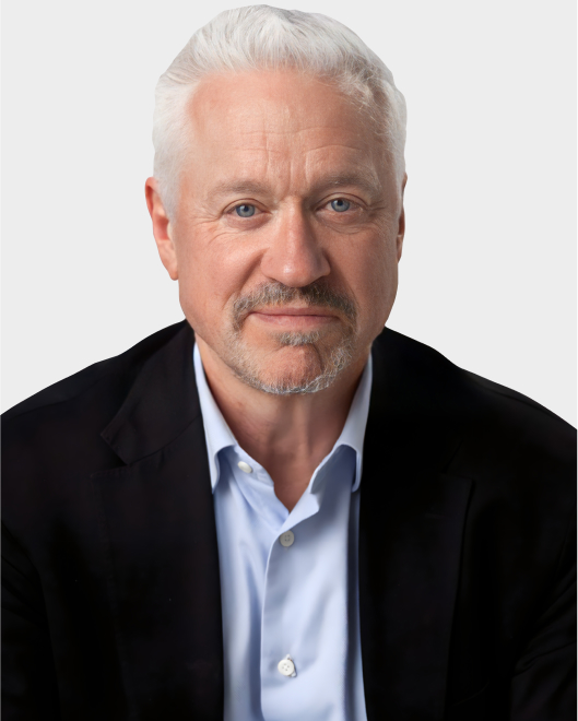A middle-aged man with short white hair, light beard, and blue eyes, wearing a black blazer over a light blue shirt, poses against a plain light background.