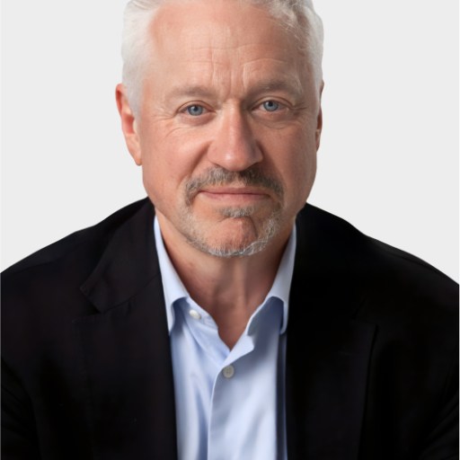 A middle-aged man with short white hair, light beard, and blue eyes, wearing a black blazer over a light blue shirt, poses against a plain light background.