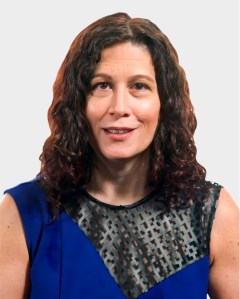 A woman with wavy brown hair wearing a sleeveless blue top with a black geometric mesh panel, posing against a plain light background.