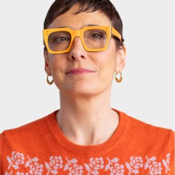 Person with short dark hair wearing large yellow glasses, gold hoop earrings, and an orange sweater with a white floral pattern, facing forward against a plain light background.