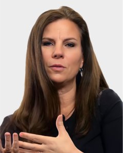 Woman with long brown hair wearing a black top, gesturing with her hand while speaking, against a plain light background.
