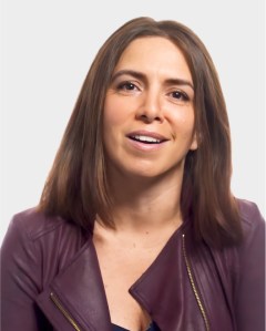 A woman with straight, shoulder-length brown hair wearing a maroon jacket sits against a plain light background, looking forward with a neutral expression.