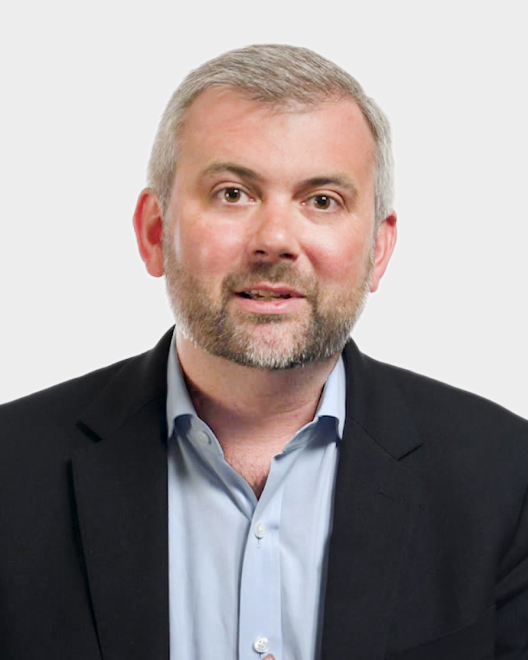 A man with short gray hair and a beard, wearing a dark blazer over a light blue shirt, poses against a plain light background.
