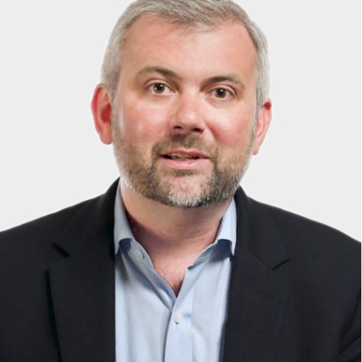 A man with short gray hair and a beard, wearing a dark blazer over a light blue shirt, poses against a plain light background.