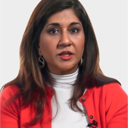 Woman with long brown hair wearing a red blazer and white top, seated against a plain light background, looking directly at the camera.