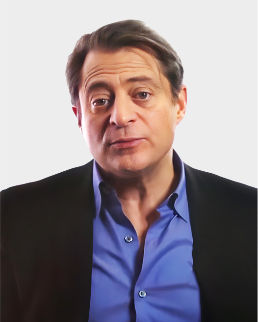 A man with short brown hair wearing a blue shirt and black blazer stands against a plain light background, looking directly at the camera with a neutral expression.