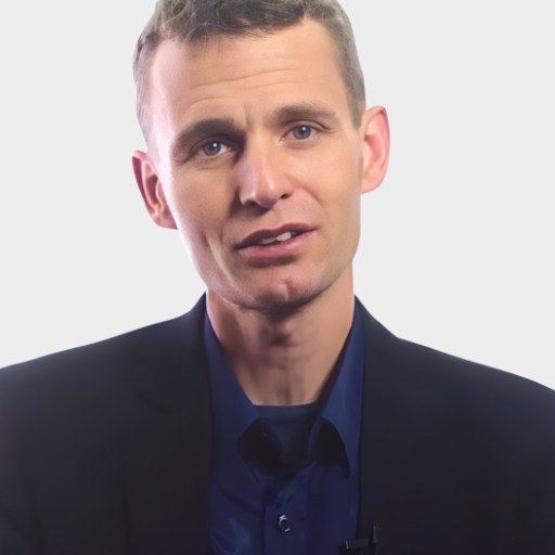A man with short light brown hair wearing a navy shirt and dark blazer faces the camera against a plain light background.