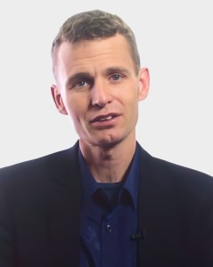 A man with short light brown hair wearing a navy shirt and dark blazer faces the camera against a plain light background.
