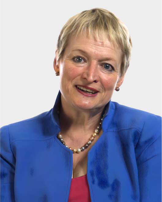 A woman with short blonde hair wearing a blue jacket, pink top, and pearl necklace, smiling at the camera against a plain light background.