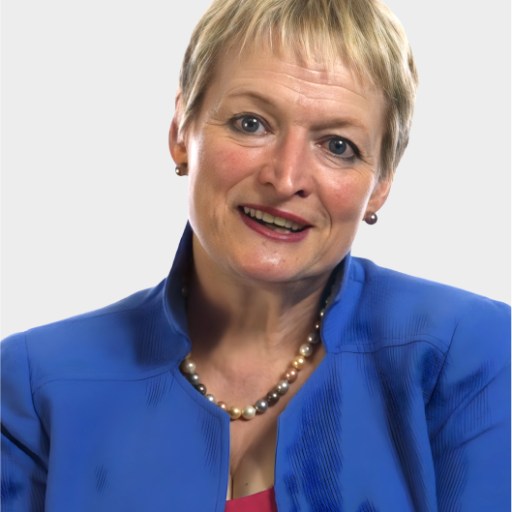 A woman with short blonde hair wearing a blue jacket, pink top, and pearl necklace, smiling at the camera against a plain light background.