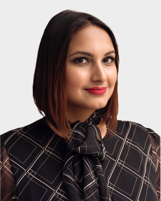 A woman with straight, shoulder-length brown hair and red lipstick, wearing a black patterned blouse with a bow, poses against a plain light background.