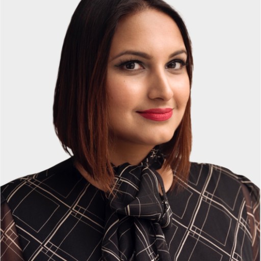 A woman with straight, shoulder-length brown hair and red lipstick, wearing a black patterned blouse with a bow, poses against a plain light background.