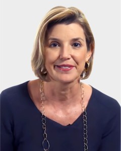 A woman with short blonde hair, wearing a dark top and a long chain necklace, is looking at the camera against a plain background.