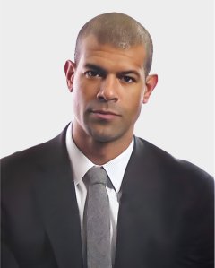 A man in a black suit and gray tie is facing forward against a plain white background.