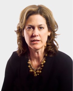 A woman with shoulder-length wavy hair wearing a black top and a gold necklace, looking forward against a plain background.