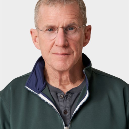 An older man with short gray hair, glasses, and a neutral expression wears a dark green zip-up jacket over a gray buttoned shirt against a plain light background.