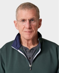 An older man with short gray hair, glasses, and a neutral expression wears a dark green zip-up jacket over a gray buttoned shirt against a plain light background.