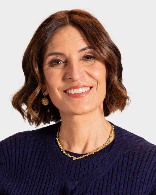 A woman with shoulder-length brown hair, wearing a navy sweater, gold necklace, and earrings, smiles at the camera against a plain light background.