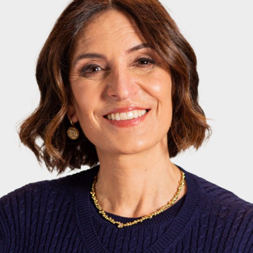 A woman with shoulder-length brown hair, wearing a navy sweater, gold necklace, and earrings, smiles at the camera against a plain light background.
