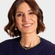 A woman with shoulder-length brown hair, wearing a navy sweater, gold necklace, and earrings, smiles at the camera against a plain light background.