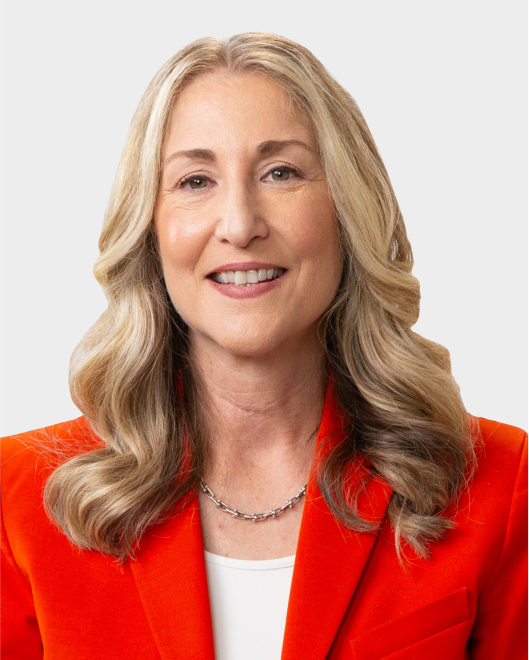 A woman with long, wavy blonde hair wearing an orange blazer, white top, and silver necklace, smiling at the camera against a plain light background.