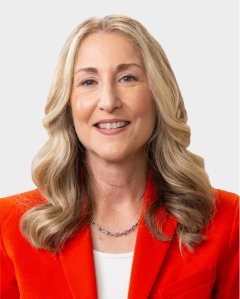 A woman with long, wavy blonde hair wearing an orange blazer, white top, and silver necklace, smiling at the camera against a plain light background.
