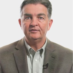 Middle-aged man wearing a gray blazer and light shirt, looking at the camera with a neutral expression, with a microphone clipped to his jacket.