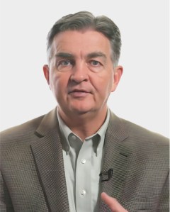 Middle-aged man wearing a gray blazer and light shirt, looking at the camera with a neutral expression, with a microphone clipped to his jacket.