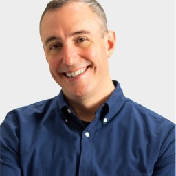 A man with short gray hair smiles at the camera. He is wearing a dark blue button-up shirt and stands against a plain light background.