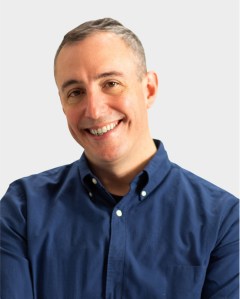 A man with short gray hair smiles at the camera. He is wearing a dark blue button-up shirt and stands against a plain light background.
