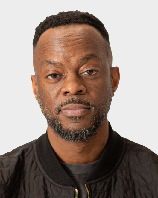 A man with short hair and a beard, wearing a black quilted jacket, looks directly at the camera against a plain light background.